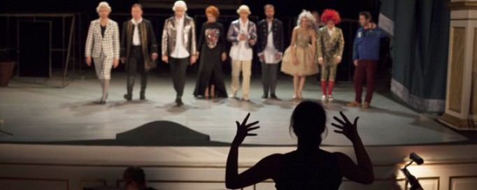 Na scenie stoją ludzie, przed sceną widać postać kobiety, odwróconej tyłem i gestykulującej.