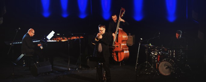 Koncert na scenie, muzycy grają na instrumentach, jeden na fortepianie, drugi na kontrabasie, a trzeci na trąbce. 