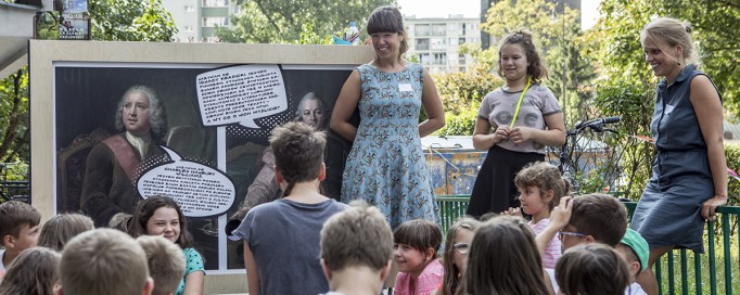 Grupa dzieci siedzi i słucha dwóch kobiet, które stoją na tle obrazu.