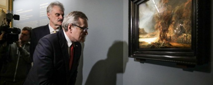 Obraz Rembrandta "Krajobraz z miłosiernym Samarytaninem" wisi na ścianie, przed nim stoi dwóch mężczyzn, którzy go oglądają.