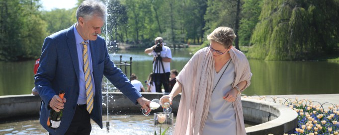 Chrzest tulipana w ogrodzie Łazienek Królewskich, mężczyzna i kobieta polewają stojący w naczyniu kwiat szampanem z butelki.