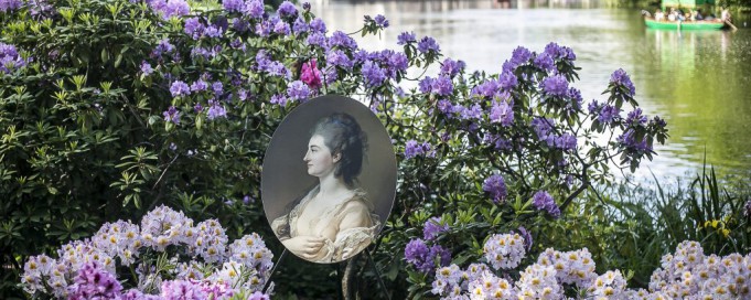 Widok na Pałac na Wyspie wśród wody i zieleni. Na pierwszym planie pośród fioletowych kwiatów wetknięta została reprodukacja portretu z kolekcji Stanisława Augusta. 