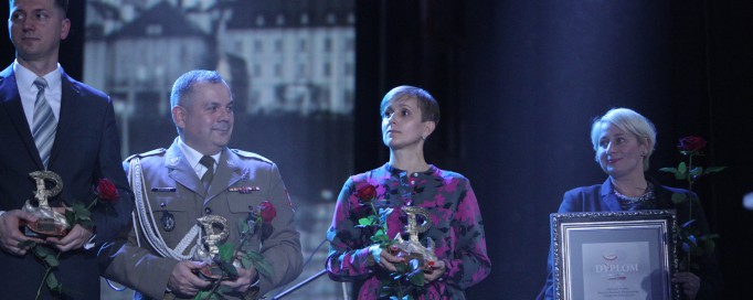 Na scenie, na tle zdjęcia fragmentu miasta, stoją dwaj mężczyźni i dwie kobiety, trzymają statuetki i róże, a ostatnia z kobiet - dyplom.