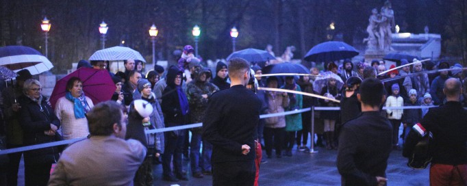 Mężczyzni z szablami stoją przed tłumem ludzi oglądających ich szermiercze umiejętności.