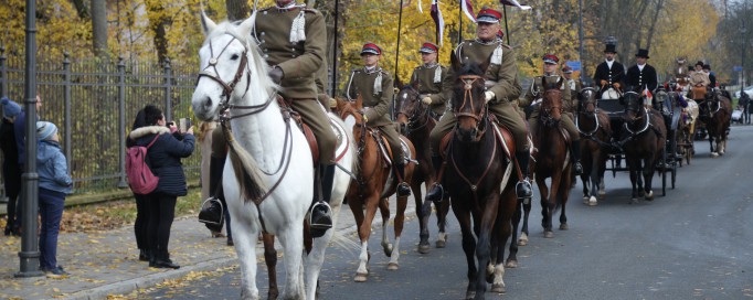 Kawalerzyści w mundurach jadą konno, z tyłu z nimi jadą dorożki. Ludzie, którzy stoją wzdłuż ulicy, robią im zdjęcia.