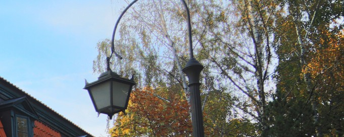 Latarnia stojąca na tle drzew i fragmentu budynku.