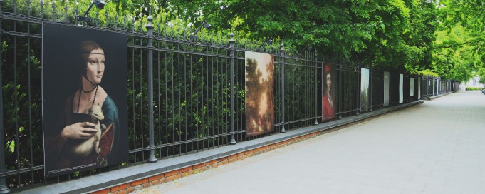 Płot galerii plenerowej, na którym wiszą plansze, na pierwszej z lewej widoczna jest kobieta trzymająca na ręku łasicę.