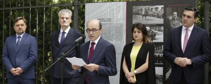 Trzech mężczyzn z lewej, kobieta i mężczyzna z prawej strony stoją pod ogrodzeniem z planszami wystawowymi, mężczyzna trzeci od lewej przemawia do mikrofonu, trzymając kartę w ręku.