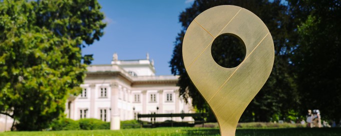 Statuetka Złota Pinezka stojąca na trawie, na tle Pałacu na Wyspie.