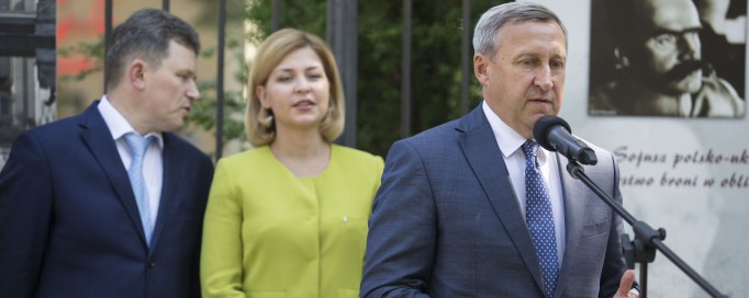 Mężczyzna w garniturze stoi przed mikrofonem, z tyłu za nim stoi kobieta z mężczyzną, widoczny jest też fragment planszy wystawowej wiszącej na płocie.