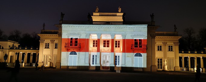 Pałac na Wyspie nocą, oświetlony iluminacją z flagą w barwach historycznej, biało-czerwono-białej flagi Białorusi.