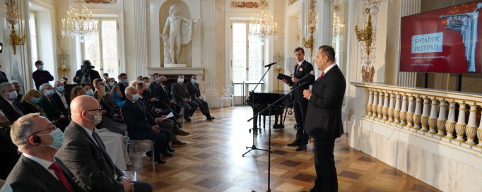 Uroczystość w Pałacu na Wyspie. W sali siedzą na krzesłach ludzie, przed ludźmi stoi mężczyzna, który mówi do ustawionego przed nim mikrofonu. 