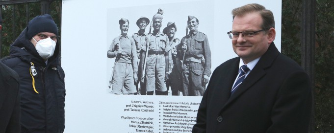 Dwóch mężczyzn stoi przy planszy wystawowej wiszącej na płocie. Plansza przedstawia grupę żołnierzy, trzymających karabiny wiszące na ich ramionach.