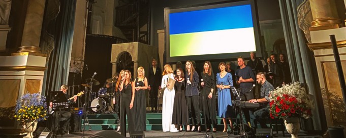 Grupa artystów stoi na scenie Teatru Królewskiego i śpiewa. Nad artystami wisi ukraińska flaga w niebiesko-żółtych barwach.