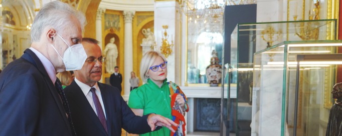 Dwóch mężczyzn w garniturach i kobieta w zielonej sukience stoją w Sali Salomona w Pałacu na Wyspie przed szklaną gablotą.