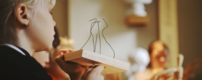 Dziewczynka z profilu trzymająca w ręku drewnianą podstawkę, do której przyczepionych jest kilka powyginanych drutów. W tle widać rzeźbione białe popiersia.