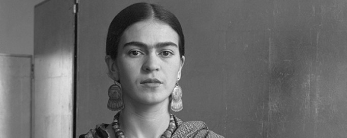 Czarno-białe, archiwalne zdjęcie Fridy Kahlo. Artystka ubrana jest w sukienkę i chustę, którą ma owinięte ramiona, oparta jest o kanapę, prawy łokieć trzyma na wiklinowym, plecionym koszyku. 