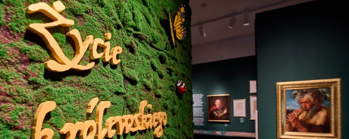 Przy wejściu na wystawę, po lewej stronie na zielonej ścianie z mchu jest napis "Życie królewskiego ogrodu". W głębi pomieszczenia widać obrazy wiszące na ścianach. 