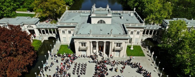 Widok z lotu ptaka na Pałac na Wyspie w otoczeniu wody i zieleni. Na tarasie przed Pałacem widać tłum ludzi.