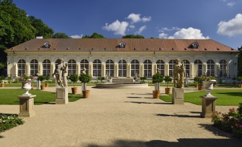 Widok na Starą Oranżerię w Łazienkach Królewskich. Przed budynkiem jest ogród z ogrodowymi alejkami i stojącymi przed nimi rzeźbami.