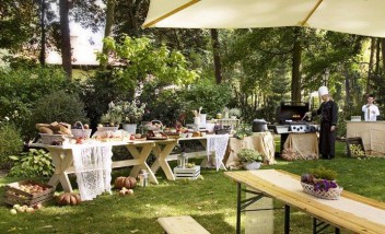 Park, w którym odbywa się piknik, stoją ławki i stoły, na których jest wystawione jedzenie.