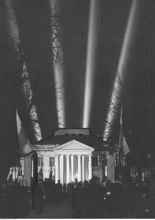 Czarno-biała fotografia. W centrum podświetlony Belweder, za nim cztery słupy światła. Przed budynkiem stoją ludzie, nad bramą wjazdową wiszą flagi.