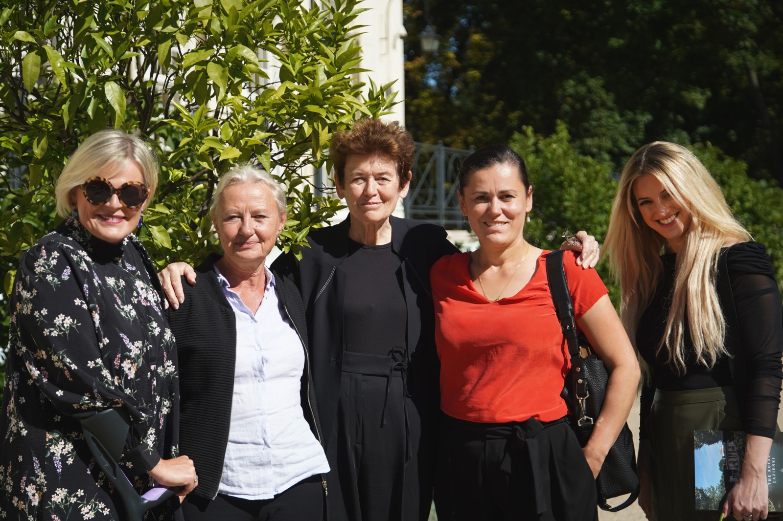Pięć kobiet stojących obok siebie i pozujących do zdjęcia w ogrodach Łazienek Królewskich, w tle za kobietami widać zieleń i fragment budynku. 
