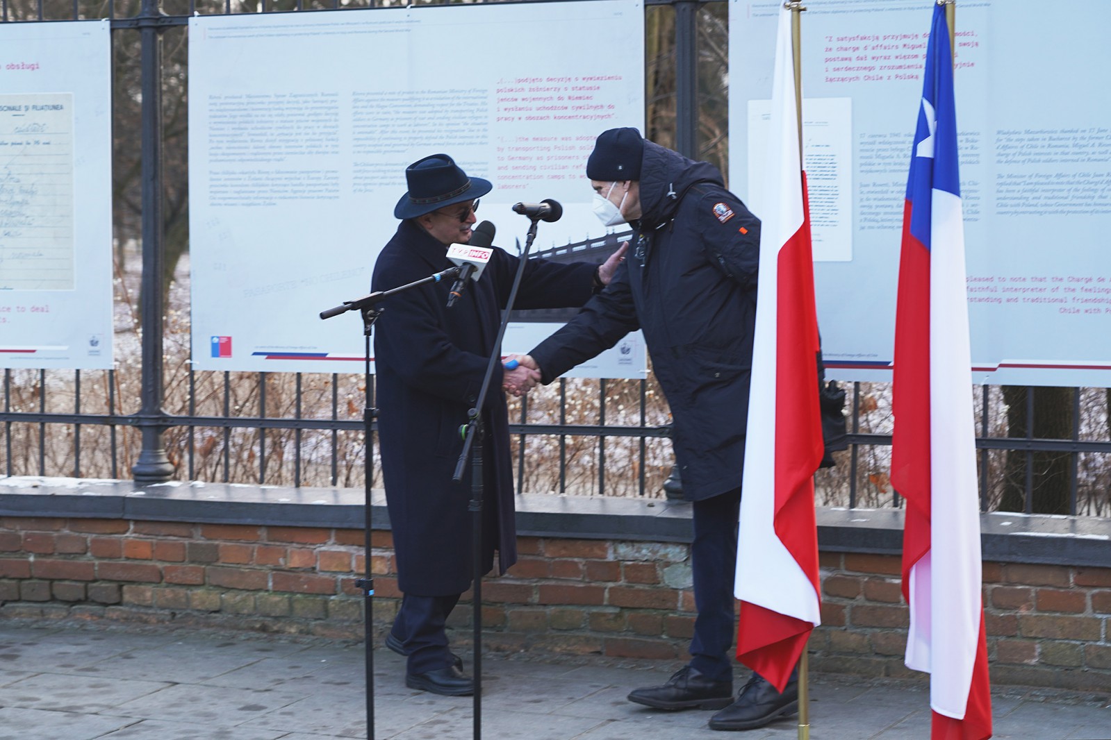 Dwaj mężczyźni w zimowych okryciach podają sobie ręce. Mężczyźni stoją na tle plansz wystawowych, wiszących na ogrodzeniu. Po prawej stronie stoją dwie flagi. 