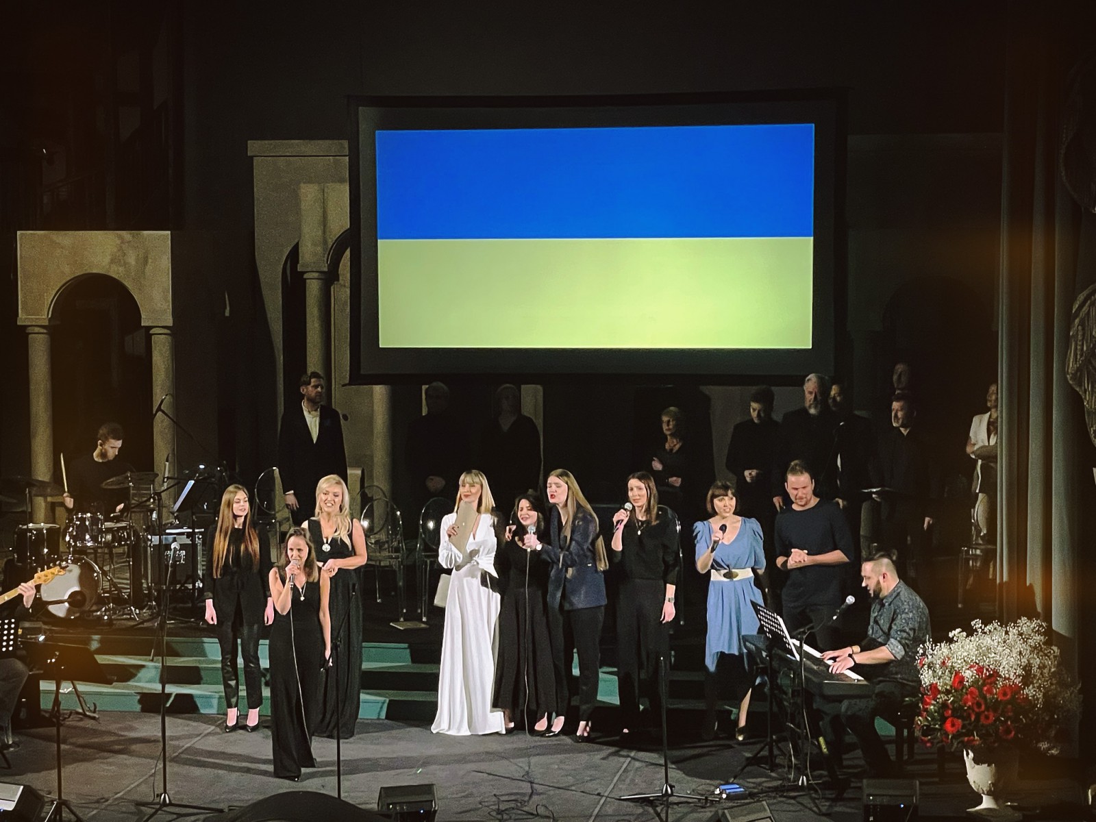 Grupa artystów stoi na scenie Teatru Królewskiego i śpiewa. Nad artystami wisi ukraińska flaga w niebiesko-żółtych barwach. 