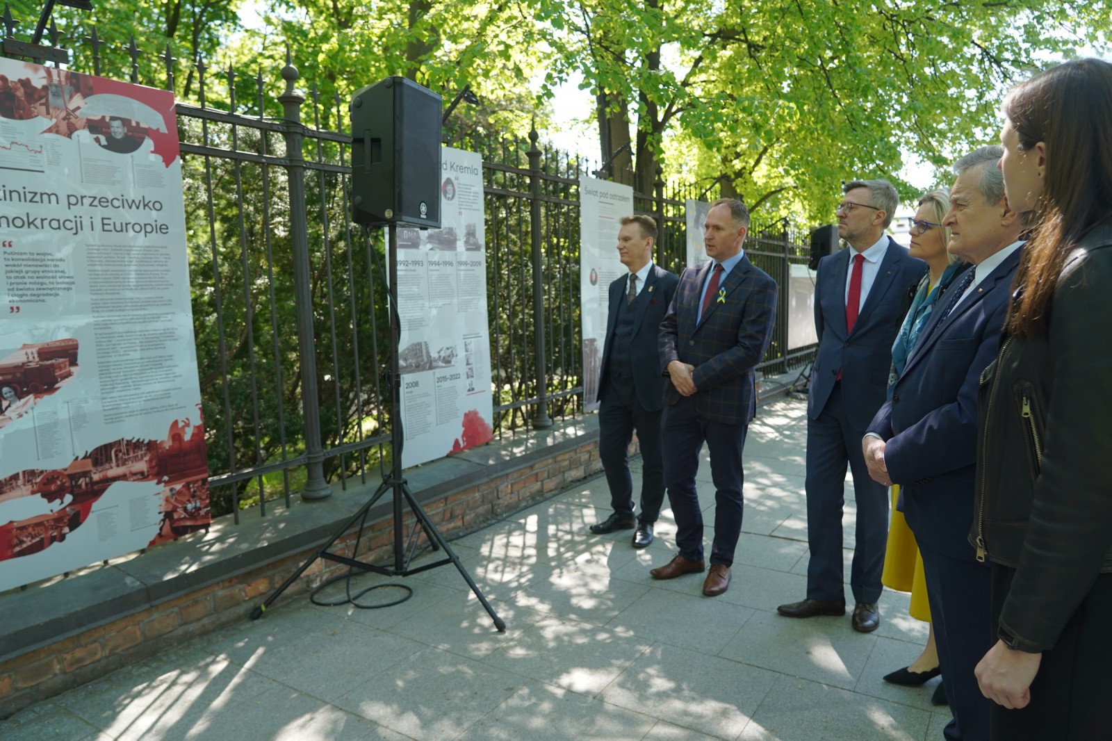 Grupa mężczyzn w garniturach i kobieta stojąca planszami wystawowymi, wiszącymi na ogrodzeniu. 