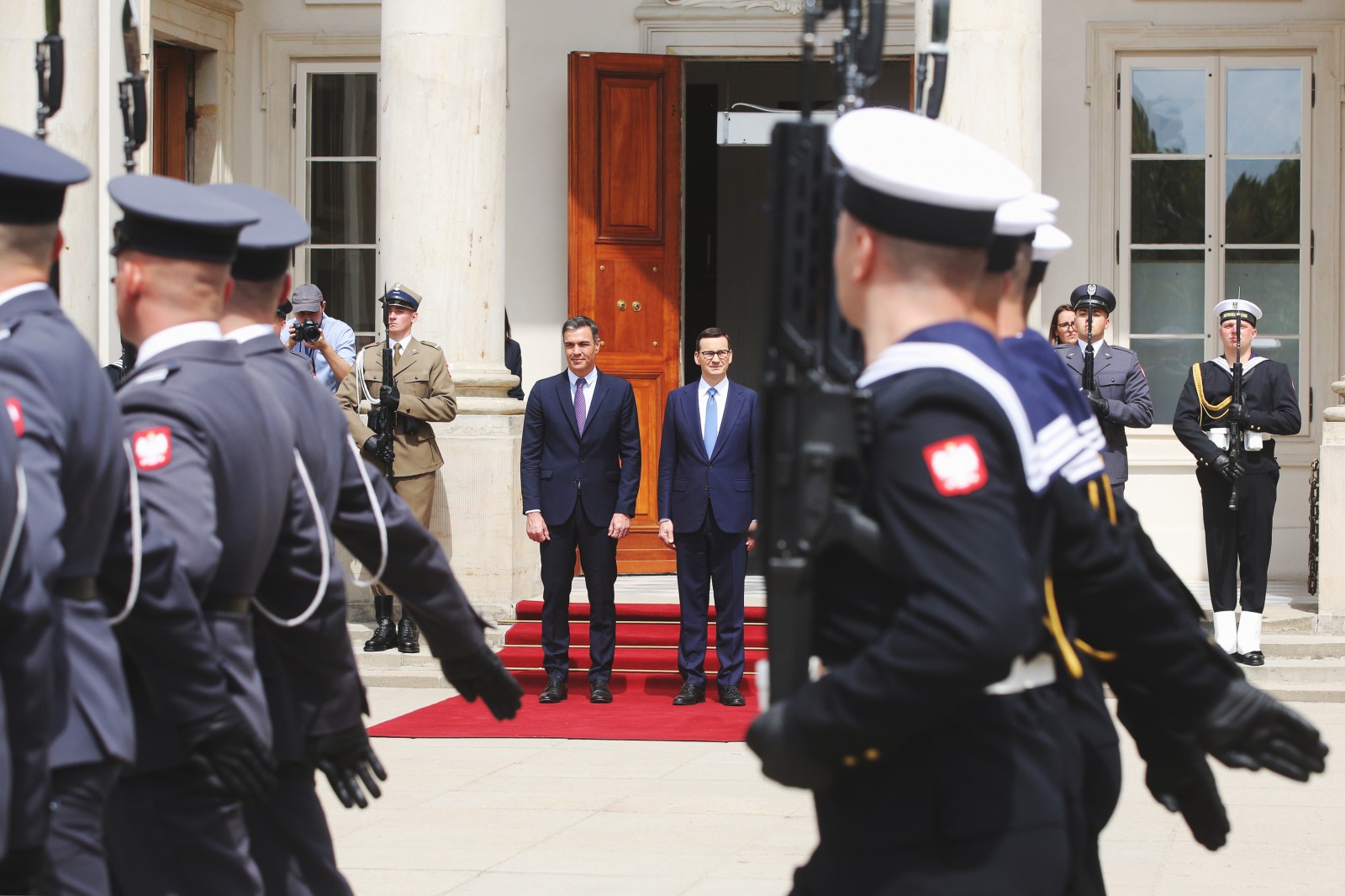 Dwóch mężczyzn w garniturach stoi na czerwonym dywanie przed wejściem do pałacu. Przed mężczyznami paradują żołnierze w mundurach, z uniesioną do góry bronią.