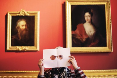 Kobieta trzyma kartkę papieru z wyciętymi otworami, którą przykłada do twarzy, w tle, za jej głową, wiszą obrazy.