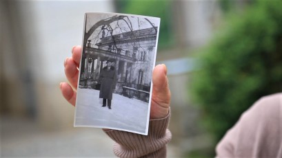 Dłoń kobiety trzymająca zdjęcie, na którym widać stojącego mężczyznę przed budynkiem. 