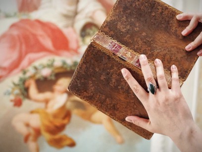 Kobiece ręce trzymające otwartą książkę na tle ściennych malowideł. 