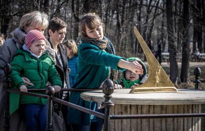 Trzy kobiety i dwoje dzieci stoją przed rzeźbą zegara słonecznego. Jedna z kobiet wskazuje ręką na zegar, który składa się z kamiennej tarczy i ustawionej na niej złotej wskazówki, uniesionej pod kątem do góry.