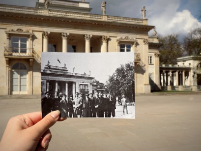 Kobieca dłoń trzymająca archiwalną fotografię na tle Pałacu na Wyspie. Na zdjęciu widoczna jest grupa ludzi pozująca na tle Pałacu na Wyspie. 