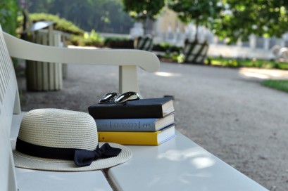 Parkowa ławka, na której leżą trzy książki, na książkach są okulary przeciwsłoneczne, a obok biały kapelusz przewiązany czarną wstążką.