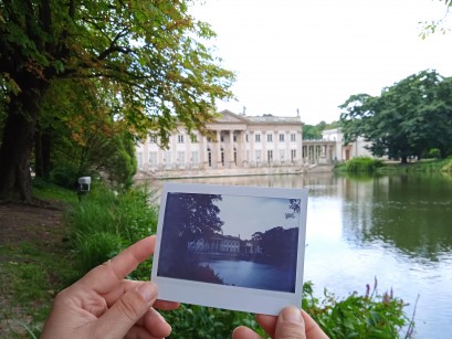 Pałac na Wyspie w otoczeniu drzew i wody. Na tle Pałacu kobiece dłonie trzymają archiwalną fotografię Pałacu na Wyspie.