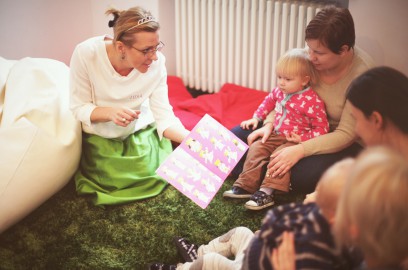 Kobieta siedzi na podłodze z otwartą książką, którą pokazuje trzem kobietom trzymającym na kolanach małe dzieci. 