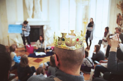Na tle grupy dzieci i dorosłych zebranych w sali Pałacu na Wyspie widoczny jest stojący tyłem mężczyzna, który ma na głowie koronę. 
