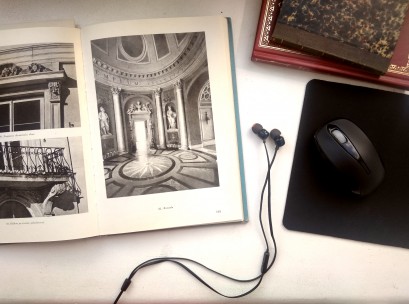 Otwarta książka ze zdjęciami obiektów Łazienek Królewskich, obok książki leżą słuchawki, podkładka z myszką oraz dwie inne, zamknięte książki. 