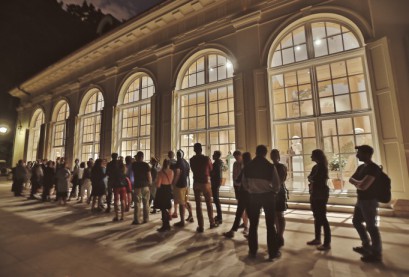 Kolejka ludzi stojąca wieczorem przed budynkiem z dużymi oknami, który jest w środku oświetlony.