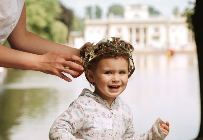 Dziewczynka z królewską koroną na głowie stoi na tle Pałacu na Wyspie. Kobiece dłonie dotykają korony na głowie dziecka.