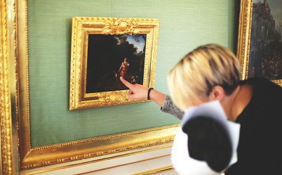 Kobieta stojąca przed obrazem z dzieckiem mającym na głowie białą, papierową koronę. Kobieta wskazuje obraz palcem. 