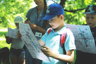 Kilkuletnie dzieci stoją w ogrodzie. W rękach trzymają mapy, na które patrzą.