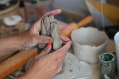 Kobiece dłonie ugniatające szary kawałek gliny nad spodkiem. Obok stoją ceramiczne kubki. 