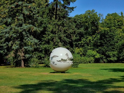 Biała rzeźba w kształcie kuli strojąca na trawie w otoczeniu drzew łazienkowskich ogrodów. 