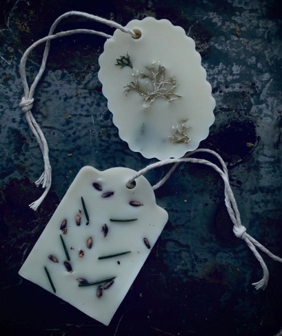 Dwie białe tabliczki z wosku, zakończone zawieszką ze sznurka, z zatopionymi w wosku kawałkami suchych roślin. 