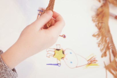 Ręka dziecka trzymająca kawałek materiału z frędzlami nad kartą z rysunkiem przedstawiającym postać. 