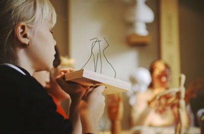 Dziewczynka z profilu trzymająca w ręku drewnianą podstawkę, do której przyczepionych jest kilka powyginanych drutów. W tle widać rzeźbione białe popiersia.